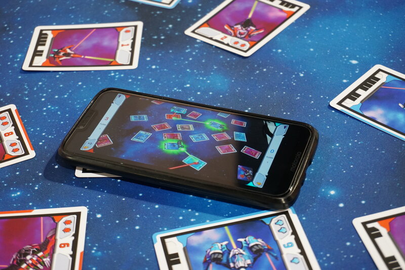 A cell phone is lying on a table and showing graphics of laser beams and explosions in augmented reality. The action on the screen is aligned with the cards on the table.